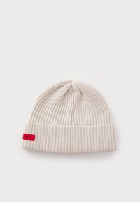 Ribbestrikket lue i lys beige, med brettkant og et lite rødt logoetikett på siden. Myk tekstur, elastisk passform.