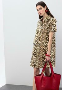 Robe chemise à imprimé léopard avec un col, des manches courtes et une coupe ample. Accessoirisée d'un grand sac à main rouge texturé et d'un bracelet en perles.