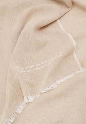 Tissu beige tissé avec bord effiloché et texte blanc pâle imprimé le long du pli dans des fibres douces et naturelles.