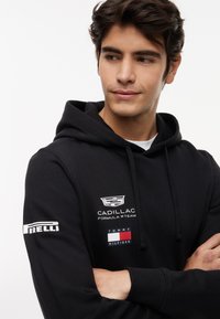 Ung mann iført svart hettegenser med Cadillac Formula 1 Team- og Tommy Hilfiger-logoer, armene i kors, ser til siden mot en hvit bakgrunn.