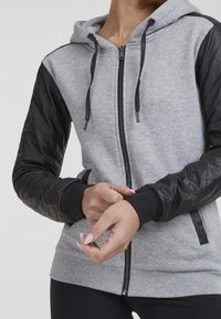 Personne portant un sweat à capuche gris zippé avec des manches en similicuir noir matelassé, les mains jointes devant sur un fond uni.