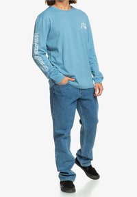 Long-sleeve blue cotton shirt with a graphic on the left sleeve, paired with light blue jeans and black sneakers, standing against a white background.