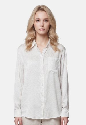 Giovane donna con capelli biondi mossi che indossa una camicia chiara a maniche lunghe con tasca sul davanti, in piedi davanti a uno sfondo semplice.