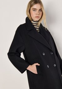 Schwarzer doppelt geknöpfter Mantel mit großen Revers, Stofftextur, zwei frontalen Taschen und sechs dunklen runden Knöpfen. Das Model trägt einen gestreiften Rollkragenpullover.