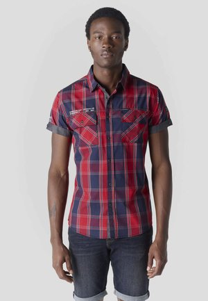 Tânăr purtând o cămașă cu mânecă scurtă, în carouri roșu și bleumarin, cu buzunare cu clapete și pantaloni scurți din denim închis, rulați.