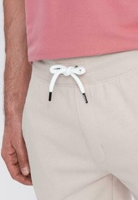Pantalones de chándal de color beige claro con un cordón blanco en la cintura, que presentan una textura suave y un ajuste relajado. Un top rosa es visible en la parte superior.