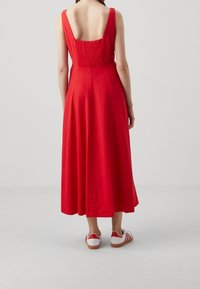 Robe longue en lin rouge avec un dos décolleté, des bretelles larges et un détail plissé. Associée à des baskets rayées blanches et rouges.