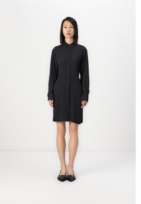 Robe chemise noire à manches longues avec fermeture à boutons, présentant un motif texturé subtil et un ourlet droit. Portée avec des ballerines noires.