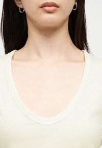 Γυναίκα που φορά μικρούς κρίκους στα αυτιά και ένα ανοιχτόχρωμο μπλουζάκι με λαιμόκοψη σε σχήμα V, εμφανίζοντας τον λαιμό, το πηγούνι και τα χείλη της σε ένα απλό φόντο.