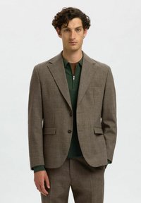 Chaqueta de traje marrón texturizada con solapas de muesca y dos bolsillos frontales, combinada con una parte superior verde con cremallera que presenta un cuello acanalado.