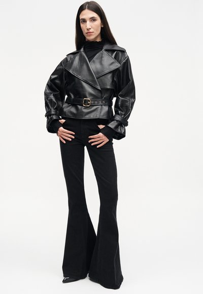 Kobieta z długimi ciemnymi włosami, ubrana w czarną skórzaną kurtkę z paskiem i czarne rozkloszowane spodnie, stoi z rękami w kieszeniach.