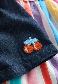 Denim fabric features a colorful embroidered bicycle design in orange and blue. The background displays striped fabric in various colors.