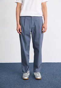 Grijze geplooide broek van lichte stof, met een losse pasvorm. Draagt bij een wit T-shirt en grijze sportschoenen.