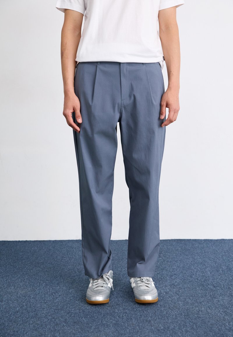 Grijze geplooide broek van lichte stof, met een losse pasvorm. Draagt bij een wit T-shirt en grijze sportschoenen.