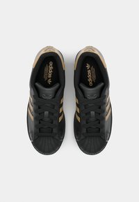 Black sneakers with three gold stripes, textured rubber toe cap, lace-up design, and an embossed logo on the tongue.