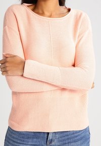 Rosa stickad tröja med runt halsband, strukturerat mönster och långa ärmar; har en ribbad kant och ribbade muddar; bärs tillsammans med denimjeans.