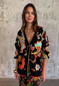 Γυναίκα που φοράει μαύρο πουλόβερ με μεγάλα πολύχρωμα λουλουδένια σχέδια, χρυσό κολιέ και φούστα που ταιριάζει, στέκεται μπροστά σε φόντο από τσιμεντένιο τοίχο.