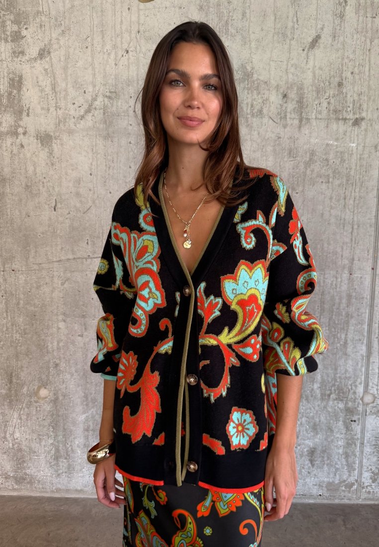 Γυναίκα που φοράει μαύρο πουλόβερ με μεγάλα πολύχρωμα λουλουδένια σχέδια, χρυσό κολιέ και φούστα που ταιριάζει, στέκεται μπροστά σε φόντο από τσιμεντένιο τοίχο.