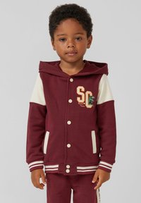 Burgundiški gobtuvų megztinis su smėlio spalvos rankovėmis, baltais sagčių uždarymais ir šoninėmis kišenėmis. Yra užrašas "SC Roar Team" siuvinėjimu ir dryžuoti rankogaliai.