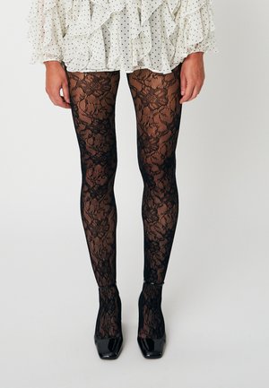 Jambes portant des collants en dentelle florale noire associés à des escarpins vernis noirs à bout carré et une jupe blanche à pois à volants.