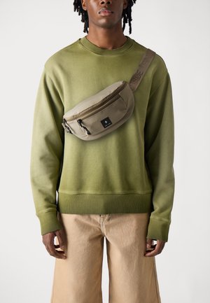 Grøn sweatshirt med ribbede manchetter, parret med beige cargo shorts. En lys brun hoftebag med lynlås og logodetalje bæres tværs over torsoen.