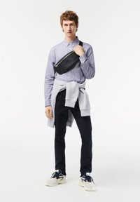 Sac en cuir noir à bandoulière, chemise à carreaux à boutons, sweat gris noué à la taille, pantalon sombre et baskets blanches avec accents bleus.