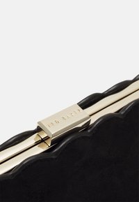 Black suede clutch with scalloped edge, featuring a gold-tone clasp with engraved brand name "TED BAKER." Elegant and structured design.