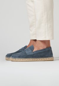 Los mocasines de ante azul presentan un detalle de lazo, una suela de yute trenzado y costuras blancas. Se combinan con pantalones de color claro.
