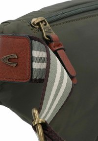 Olijfgroene tas met een gestructureerde bruine schouderriem, een grijs-witte gestreepte accenten en een bronzen rits. Voorzien van metalen hardware en stikseldetails.