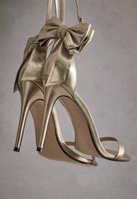 Korkeakorkoiset sandaalit metallinhohtoisessa kullassa, joissa on suuri olanvaatekoriste, avonaiset varpaat ja stiletto-korot; sileä pinta ja minimalistiset hihnat.