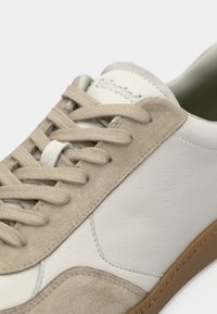 Witte en beige sneaker met suède en leren materialen, platte veters, ronde neus en lichte zool. Bevat een subtiele merklogo op de tong.
