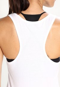 Haut blanc sans manches avec un dos nageur, comprenant un soutien-gorge de sport en mesh noir en dessous. Texture douce et emmanchures ouvertes pour le confort.