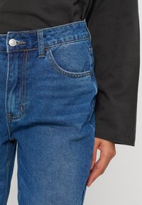 Personne portant un jean bleu en denim et une chemise noire à manches longues, la main droite détendue à côté.