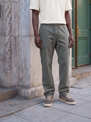 Homme portant un pantalon cargo gris, des baskets beiges et un t-shirt blanc, debout sur un trottoir en pierre près d’un mur en pierre sculpté et d’une porte verte.