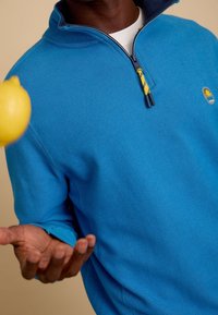 Blauwe pullover met een half-rits ontwerp, gestructureerde gebreide stof en een gele ritstrekkertje. Heeft een klein logo op de borst.