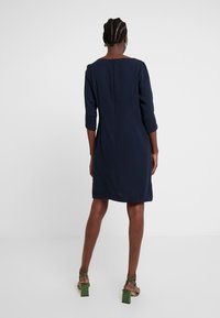 Navyblaues Kleid mit Dreiviertelärmeln, geradem Schnitt und Reißverschluss hinten. Tragen mit grünen, hochhackigen Sandalen im Schlangenmuster.