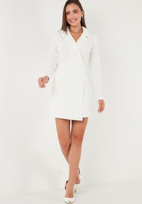 Woman standing, wearing a white long-sleeve blazer dress with lapels, paired with white pointed-toe heels, smiling against a plain background.