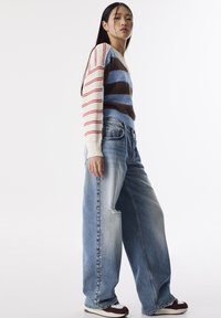 Maglione a maglia con strisce rosse, blu e marroni, abbinato a jeans a gamba larga di un leggero blu scolorito, con dettagli di bottoni lungo il lato.