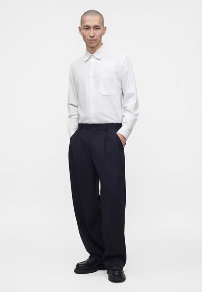 Homme portant une chemise blanche boutonnée, un pantalon noir ample et des chaussures noires, debout les mains dans les poches sur fond blanc.