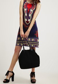 Žena v barevných vzorovaných šatech bez rukávů, držící velkou černou kabelku, v černých sandálech na vysokém podpatku s třásněmi.