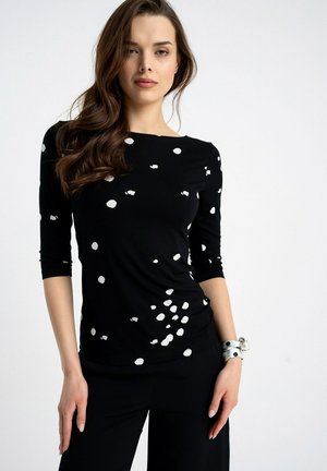 Kobieta w czarnej bluzce z długim rękawem w rozproszone białe groszki, czarnych spodniach i srebrną bransoletką, stojąca na białym tle.