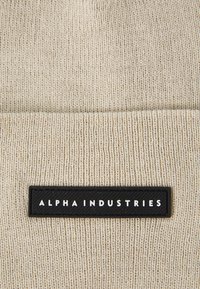 Gorro bege de malha com um retângulo preto apresentando o texto "ALPHA INDUSTRIES" em branco, exibindo padrões texturizados.
