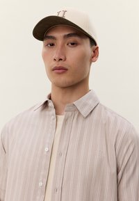 Ung mann iført en beige stripete skjorte med knapper og beige baseballcaps med en minimalistisk logo, vendt mot kamera med et nøytralt uttrykk.