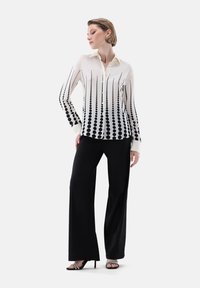 Camicia bianca con pattern a punti neri in gradiente, abbinata a pantaloni neri a gamba larga e sandali con cinturino. Tessuto morbido, vestibilità su misura.