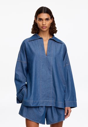 Femme aux cheveux bouclés porte une chemise en jean bleu ample à manches longues avec col ouvert et un short en jean assorti, debout sur un fond blanc.