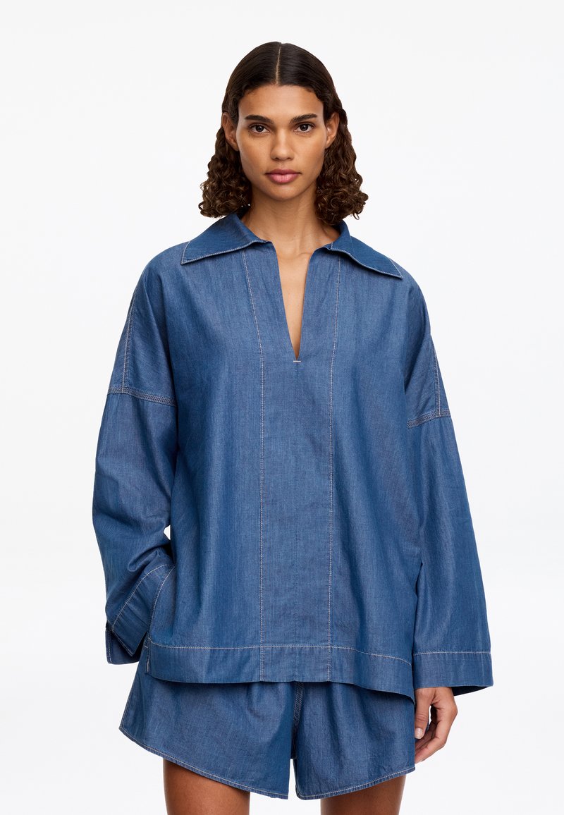 Femme aux cheveux bouclés porte une chemise en jean bleu ample à manches longues avec col ouvert et un short en jean assorti, debout sur un fond blanc.
