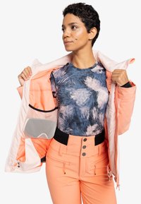 Lichtroze jack met een geborduurd interieur en een mesh pocket, gelaagd over een blauw-wit patroon top en oranje skibroek met knopen.