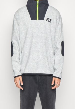 Jasnoszara bluza z polaru z czarnym kołnierzem i łokciowymi łatami, neonowo żółtym zamkiem błyskawicznym i małym logo New Balance na przedniej części klatki piersiowej.