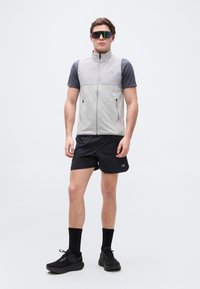 Moški, oblečen v sivo brezrokavnik z zadrgo, temno sivo kratko majico, črne kratke hlače, črne nogavice, črne superge in velika odsevna sončna očala.