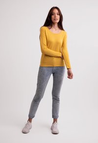 Gelbes Langarmoberteil mit Rundhalsausschnitt, kombiniert mit hellblauen Slim-Fit-Jeans und weißen Freizeitschuhen. Schlichte Textur, lässiges Design.